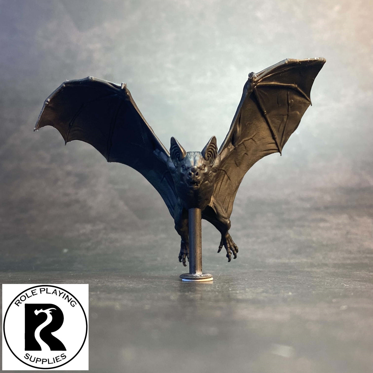 A detailed dnd miniature bat figurine with wings spread, mounted on a pole, set against a smoky, atmospheric background. A logo for 'Role Playing Supplies' is visible in the bottom-left corner.